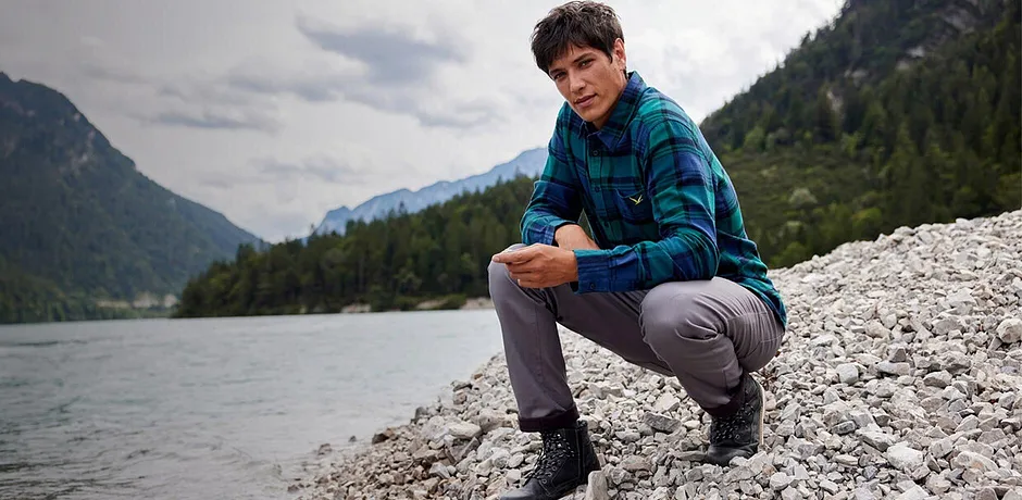 Un randonneur vêtu d'une chemise à carreaux et d'un pantalon gris assis au bord d'une rivière dans un paysage de montagne.