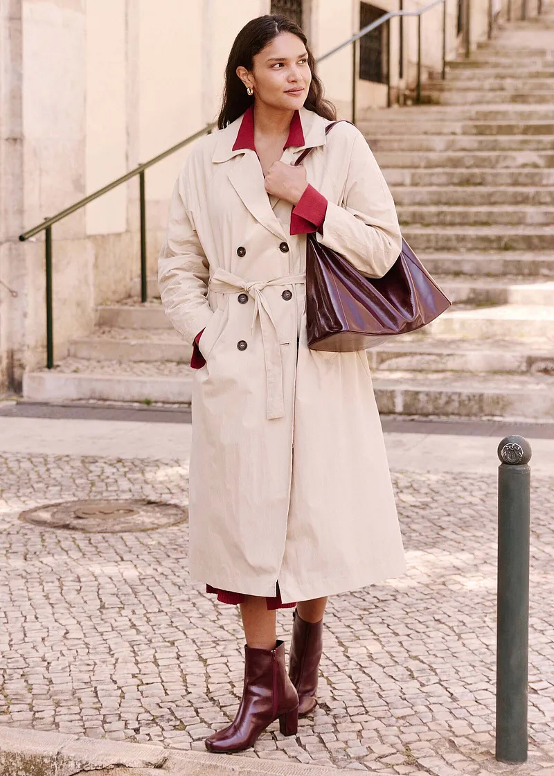 Manteau élégant couleur crème avec ceinture, chemisier rouge et bottes bordeaux. Un sac à main chic complète le look.