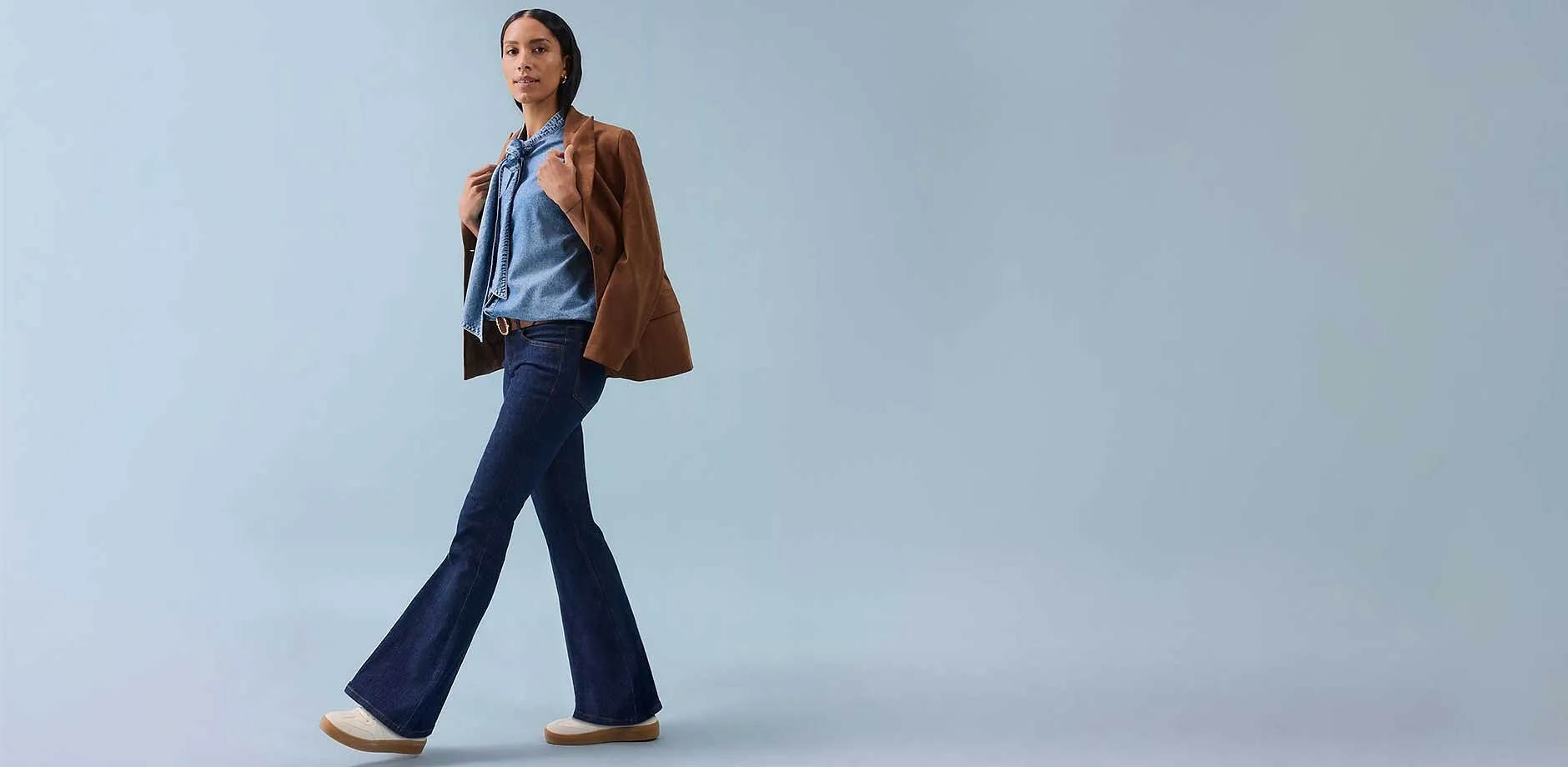 Une femme en pantalon à patte d'eph, top sans manches et veste marron portée nonchalamment sur les épaules.