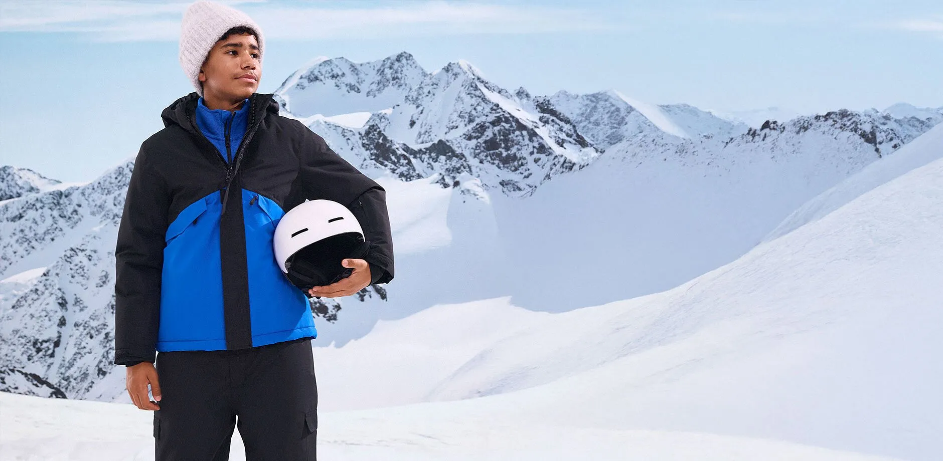 Un garçon vêtu d'une veste de ski bleu et noire tenant dans les mains un casque devant des montagnes enneigées.