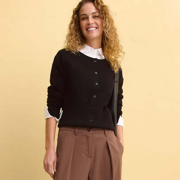 Gilet en maille noir avec boutons sur un chemisier blanc, associ&eacute; &agrave; un pantalon marron.