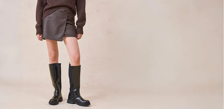 Jupe enduite associée à des bottes hautes jusqu'au genou et un pull marron.