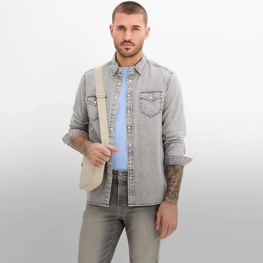 Chemise en jean claire sur un T-shirt bleu clair, compl&eacute;t&eacute;e d'un jean gris et d'une besace beige.