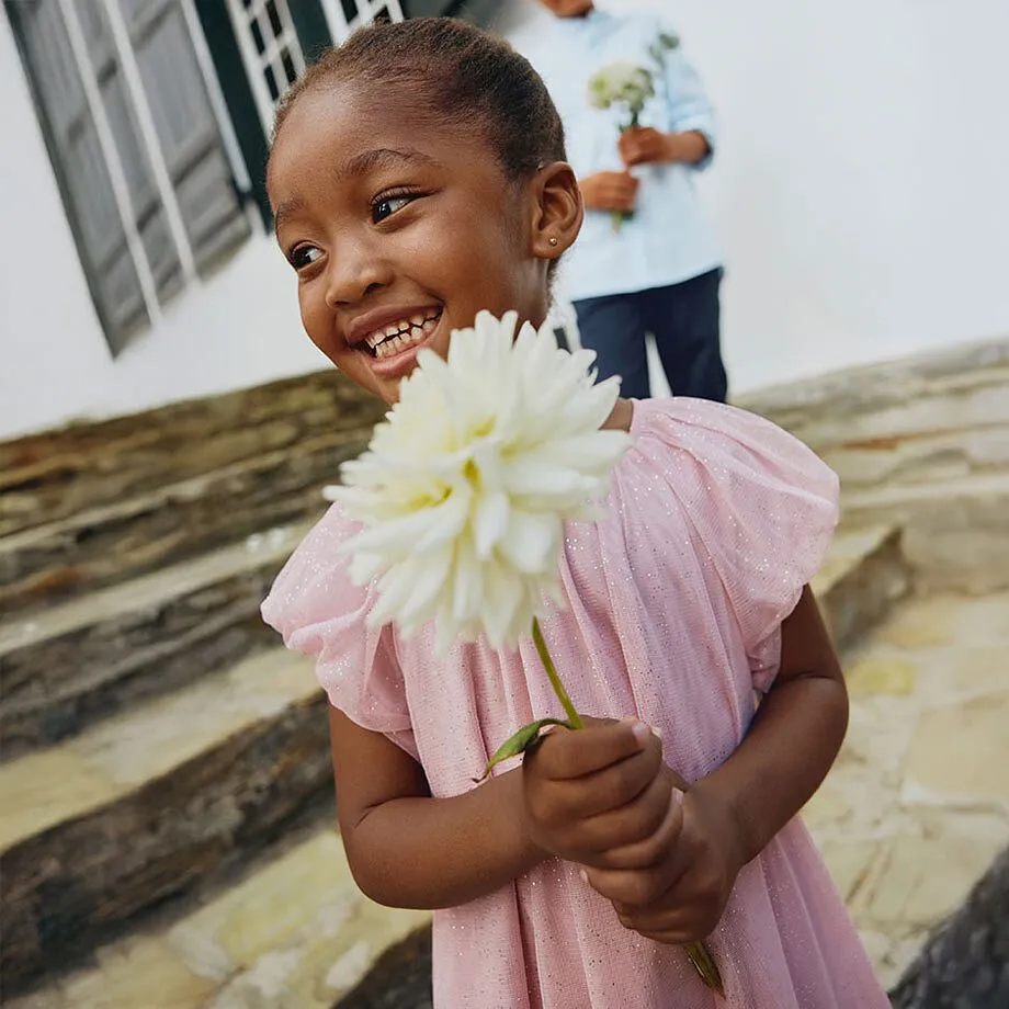 Enfant radieuse v&ecirc;tue d'une robe rose avec manches &agrave; ruch&eacute;s, tenant une fleur blanche.