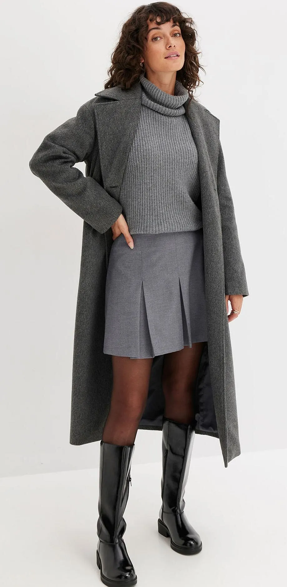 Manteau gris en laine, pull &agrave; col roul&eacute;, jupe pliss&eacute;e et bottes noires.